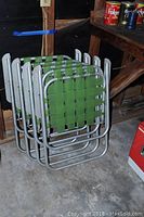 Stack of four green plastic webbing folding lawn chairs with metal frames folded and stacked vertically on a concrete floor.