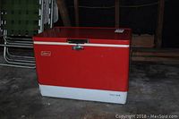 Vintage red and white Coleman cooler with 'Trey Big' label, closed lid, metal latch and side handles, showing signs of wear and storage dust.