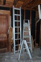 Overview of three aluminum ladders (step ladder, standard ladder, extension ladder) with coil of rope hanging on one ladder, in storage area.