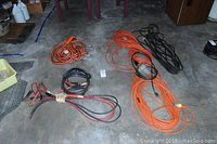 Photo of assorted garage items including jumper cables, heavy duty extension cords in orange and black, and rope cables laid out on concrete floor.