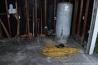 Photo showing wall with multiple garden tools including rake, shovel, hoe, post hole digger, and water heater tank in corner with extension cords on floor.