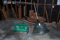 Wide photo showing rusty wheelbarrow, green push spreader, and handheld leaf rake in storage area