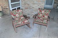 Photo showing both wooden chairs with floral cushions, placed outdoors on a concrete patio near a brick wall.