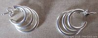 Pair of small sterling silver triple hoop earrings laid flat on a fabric background showing front and back sides including post backs.