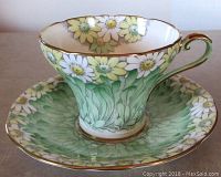 Side view of the cup sitting on the saucer showing the green background with white and yellow daisies and gilded edges.