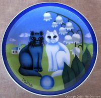 Top-down photo of the round porcelain plaque showing two cats, lily of the valley flowers, houses in background, and ball. Colors mainly blue, white, green.