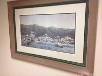 Photo of a framed wall art print showing a coastal town with boats on water, surrounded by hills, and a wooden frame with a green matte.