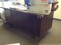 Photo showing side view of dark wooden office desk with a box on top, highlighting the repair needed at the base and the desk's structure.