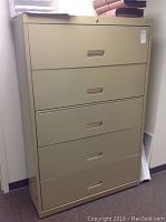 Front view of beige metal filing cabinet showing all 5 drawers with recessed handles and label slots. Cabinet stands tall with slight wear visible on the finish.