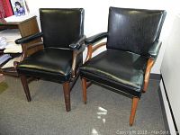 Two black leather armchairs with wooden frames placed side by side in office setting. Shows general wear and use.