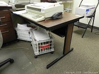 Side-angle photo of the office table showing the wood grain top, black metal frame, and shelf underneath, with some wear visible on edges. Items and paper crates stored underneath.