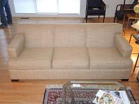 Front view of the tan fabric couch showing three seat cushions and short wooden legs, placed on hardwood floor by a window.