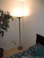 Full view of the silver-colored torchiere floor lamp standing next to a bed and a plant, plugged into a wall outlet.