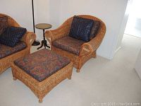 Two wicker chairs, one visible with paisley pattern cushions and pillows, and wicker ottoman with matching cushion in a bright carpeted room.
