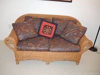 Front view of wicker loveseat with patterned cushions and multiple pillows including a red decorative pillow (not included).