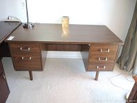 Front view of wooden office desk showing six drawers and metal handles, standing on four legs, some light scratches visible on surface.