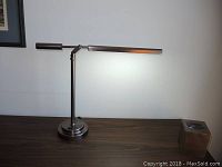 Full view of metallic desk lamp turned on on wooden surface with visible framed artwork in background.