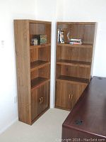 Pair of wood grain MDF bookshelves in corner of room, each shelf has three open shelves and closed cabinet lower section.