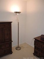 Full view of metallic torchiere floor lamp standing at corner between two wooden furniture pieces, plugged in but untested.