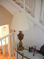 Brass finish table lamp with pleated off-white lampshade sitting on glass table near stairwell, showing lamp turned off.