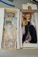 Two boxed and wrapped musical dolls lying side by side, showing one doll with blonde hair and elaborate navy blue dress, the other wrapped in bubble wrap in its box.