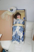 Full view of the doll in original box, showing the blue satin dress with white lace details and bonnet attached to the box.