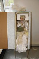 Porcelain doll Angelica lying in an open box, partially wrapped in plastic and foam padding, showing the detailed gown and hair.