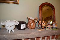 Shelf display with footed milk glass bowl, dark pottery bowl, Staffordshire pitcher, arched wood framed mirror, and four Royal Crown Hummel figurines.