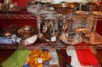 Shelf view of multiple silver plated items including trays, pitchers, serving bowls, and utensils, showing variety and quantity of lot