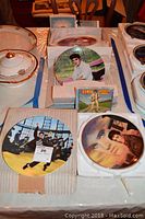 Photo showing four Elvis collectible plates arranged on a table with the CD and boxes, showcasing different images of Elvis Presley.