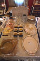 Overview of vintage dishware set on table including platters, bowls, gravy pitcher, champagne flutes, custard cups, warming dish, and cutting boards.