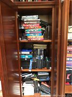 Photo showing wooden cabinet with multiple stacks of DVDs and VHS tapes inside, some cases black and some colored.