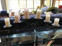 Full view of black metal candle stand with six cream pillar candles, shown on glass surface in front of a couch with visible indoor setting.