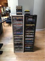 Front view of two DVD storage racks, one white and one black, filled with DVDs and topped with stacks of 8-track tapes.