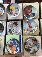 Six limited edition collectible plates shown in their original boxes, featuring various detailed illustrations of people, including children and adults, in vintage and pastoral settings.