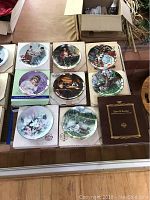 Nine collectible plates with boxes laid out on a table, including floral, animal, and historical scenes.