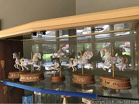 Wide photo showing all five carousel music boxes on a glass shelf, highlighting sizes and uniform design with carousel horses mounted on wooden bases.