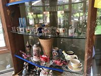 Display cabinet showing assortment of beer steins, Coca Cola art pieces made from cans, ceramic cup, and decorative miniatures