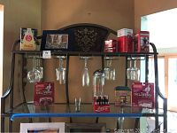 Shelf display of Coca-Cola collectibles including dispensers, glasses, tins, matted art and delivery vehicle packaging while also showing wine stemware hanging from the rack.