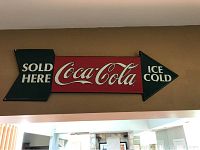Rectangular metal Coca-Cola arrow sign stating 'Sold Here Ice Cold' in red, white, green colors mounted on wall.