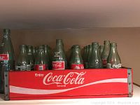 Red wooden Coca-Cola crate filled with empty and partially filled glass Coca-Cola bottles of various styles, showing embossed and printed logos.