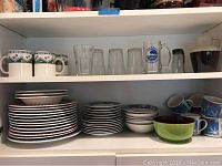 Full shot of shelves showing stacked Coca-Cola branded plates, bowls, mugs, clear glasses and decorative cups.