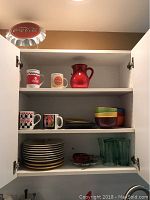 Open cabinet with various Coca-Cola mugs, red pitcher, stacked plates, Mikasa bowls, green glasses, and smaller dishware.