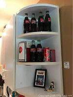 Overall view of a white shelf holding four large Coke glass bottles, three smaller bottles, a Coke can, a napkin dispenser, a framed ad and a snowman figurine.