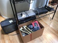 Metal shelving unit with Samsung DVD player, Sony VHS recorder, and other electronics. Box with cables and manuals on the floor.