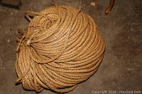 Full view of a large coil of thick, brown natural fiber rope with a braided texture, laid on a rough floor.