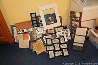 Photo showing a wide variety of photo frames arranged on floor against yellow wall and wooden door, includes a large white frame, several black frames, and other assorted sizes and finishes.