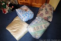 Five comforters of various colors and patterns on floor in front of wooden cabinet. One comforter is packaged with blue snowflake pattern, another packaged with blue plaid pattern, and three unpackaged comforters including a beige solid and floral pattern.