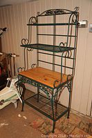 Full view of the metal bakers rack showing green metal frame with wood middle shelf and decorative curved sides.