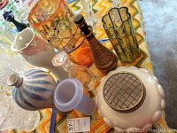 Photo showing an assembly of glass and ceramic vases and decanters on a patterned tablecloth, including the iridescent bowl vase with brass top and various other decorated vases in multiple colors and styles.